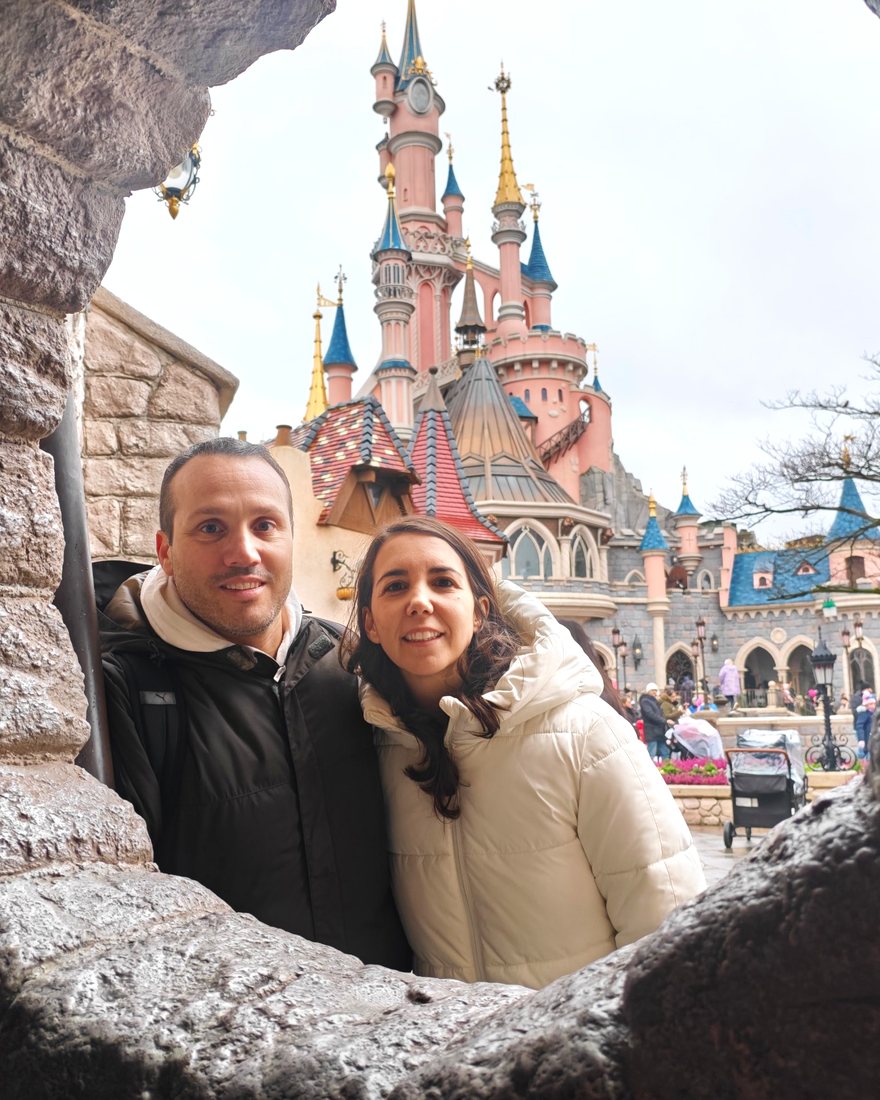 Pareja sonriente en el parque, asomada entre rocas con torres rosas al fondo